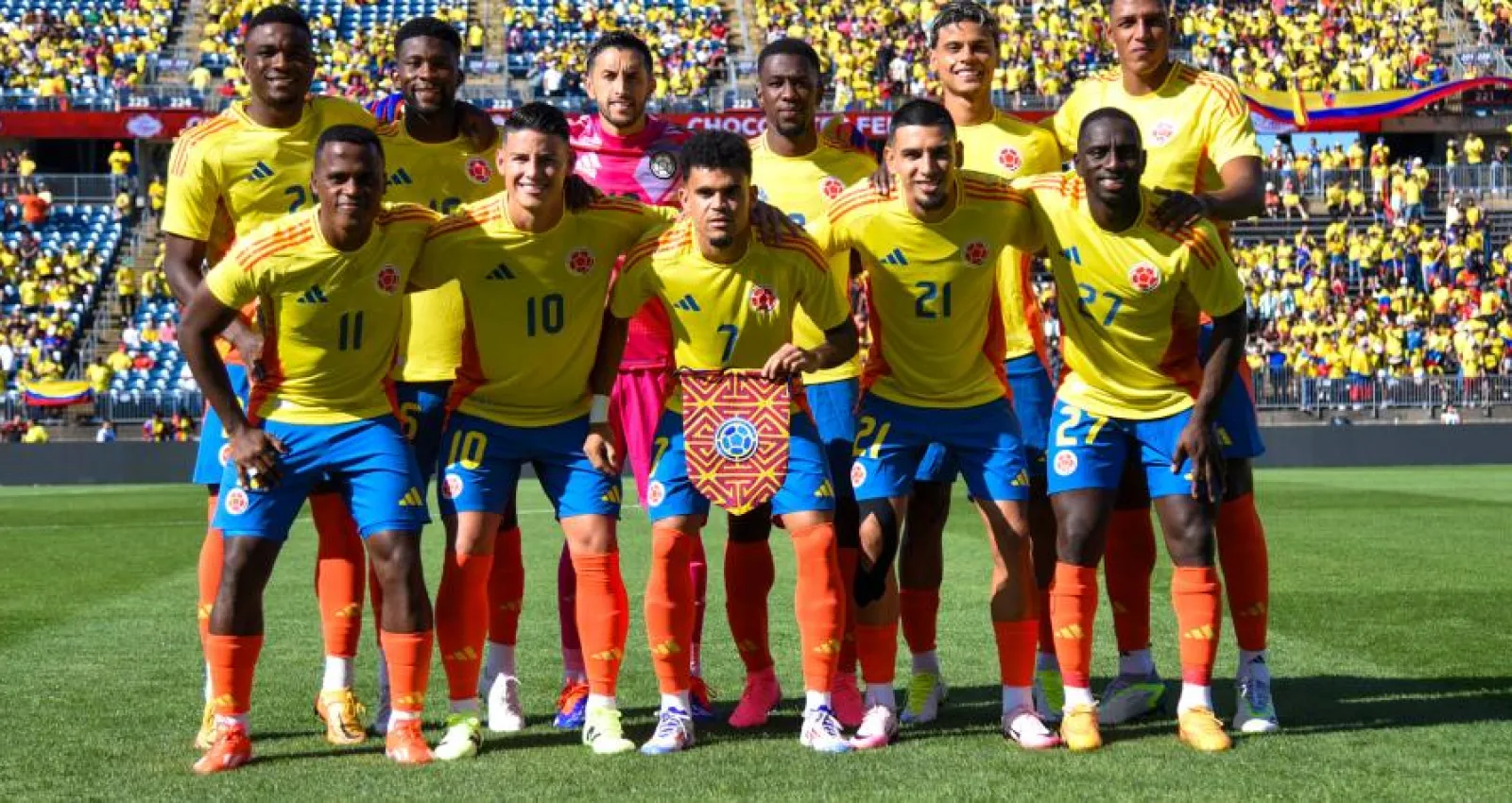 Foto de un equipo de fútbol tomados de los brazos con un fondo muticolor