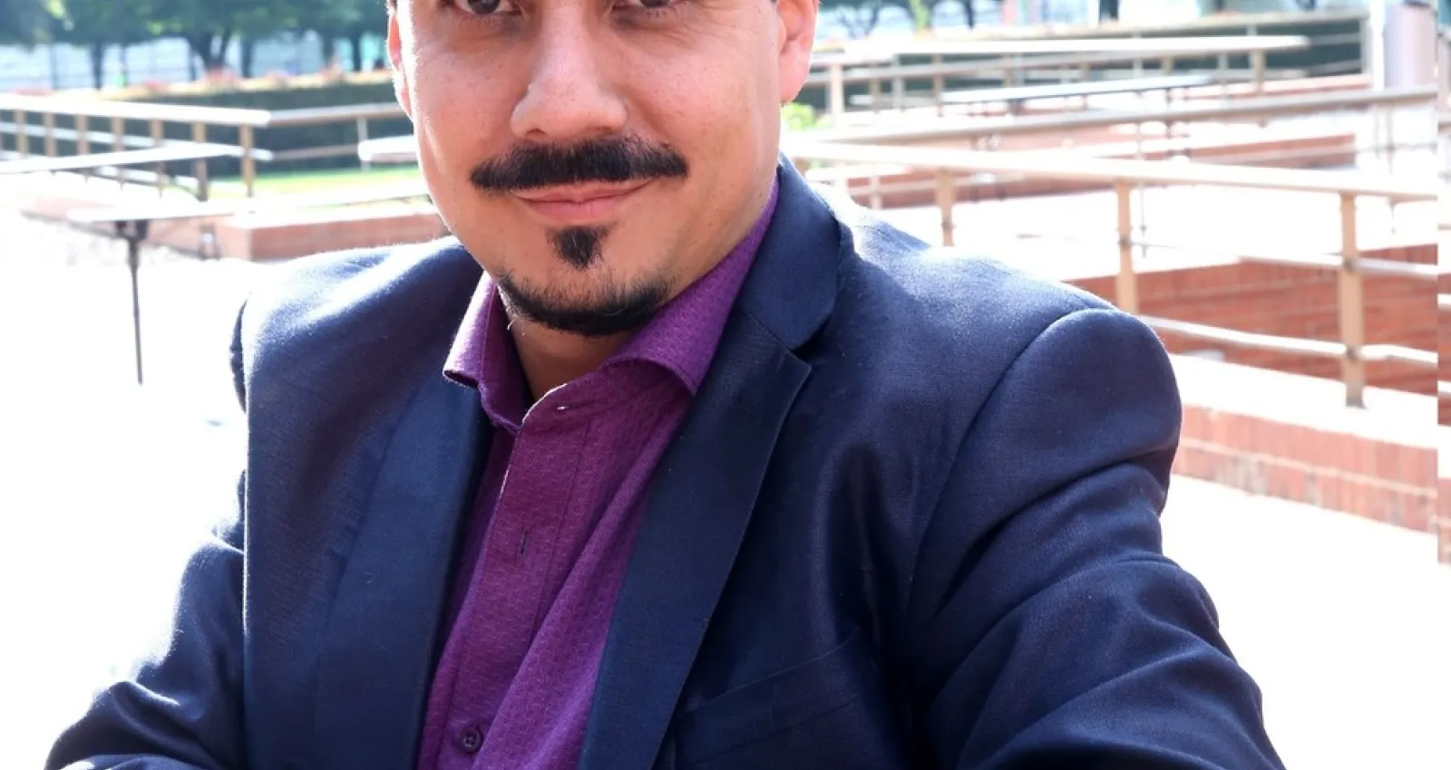 Foto de un hombre blanco con bigote mirando a la cámara, utiliza blazer azul y camiseta vinotinto