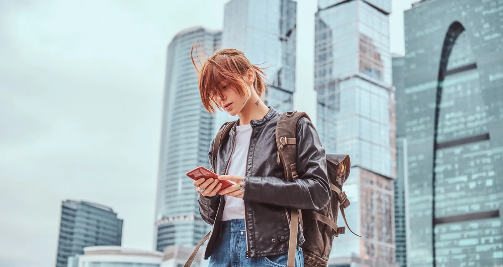 Foto de una mujer con chaqueta roja mirando un celular y unos edificios de fondo
