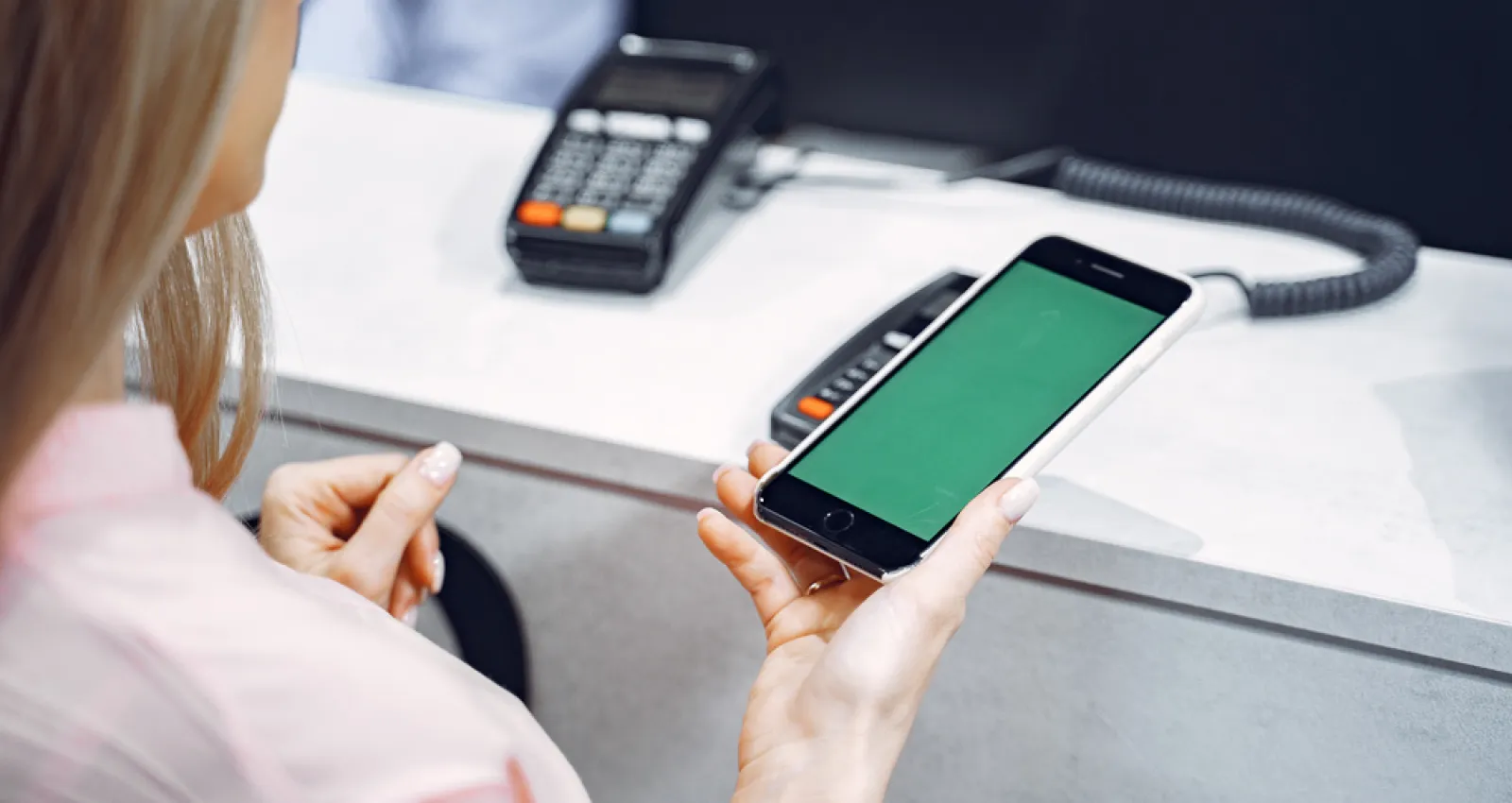 Foto de una mujer portando un celular en la mano con un datafono al fondo