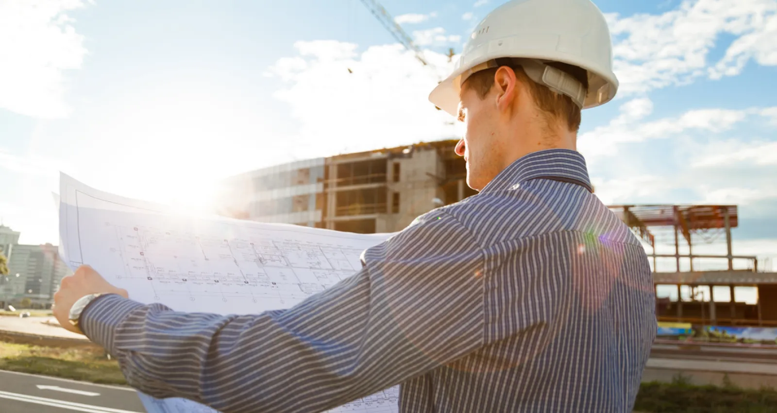 Fotografía de un constructor con camiseta azul sosteniendo en sus manos un plano de construcción y una edificación de fondo