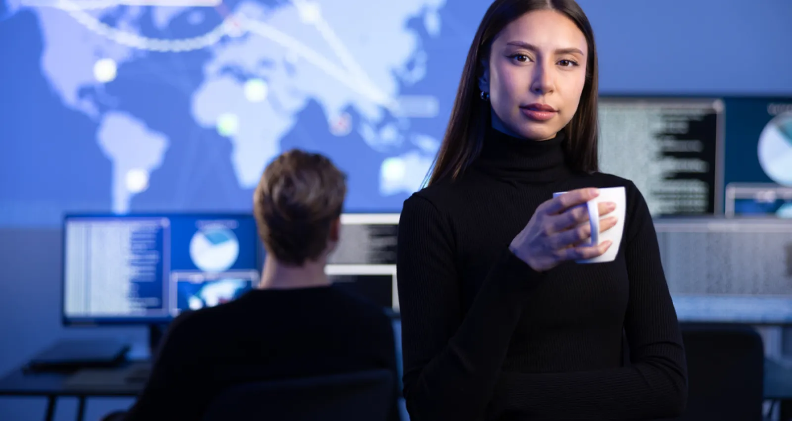 Mujer mirando a la cámara con un fondo de una persona trabajando en un computador y un diseño de continentes arriba de él