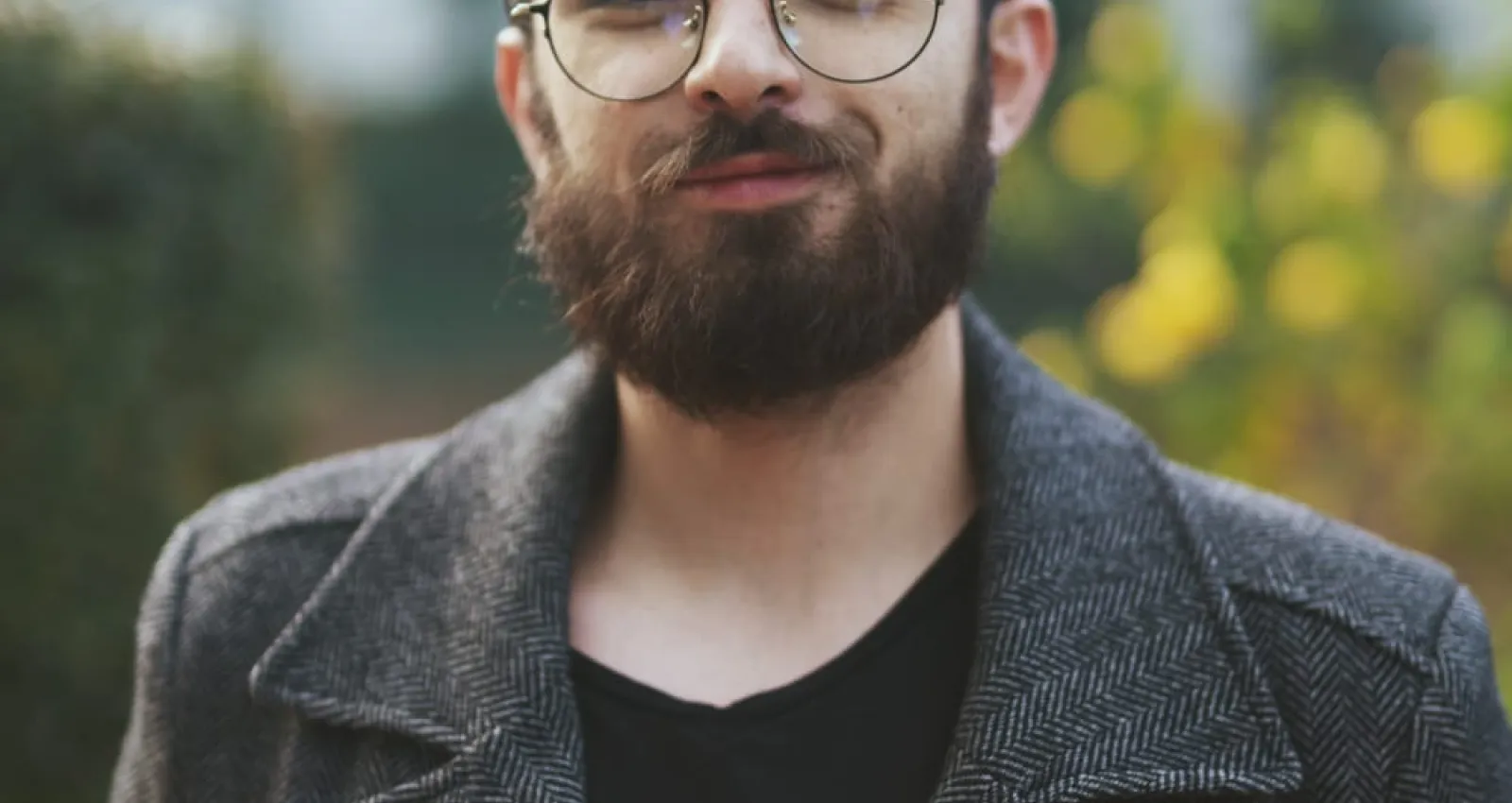 Foto de un hombre con barba y fondo verde con una gabardina gris mirando la cámara