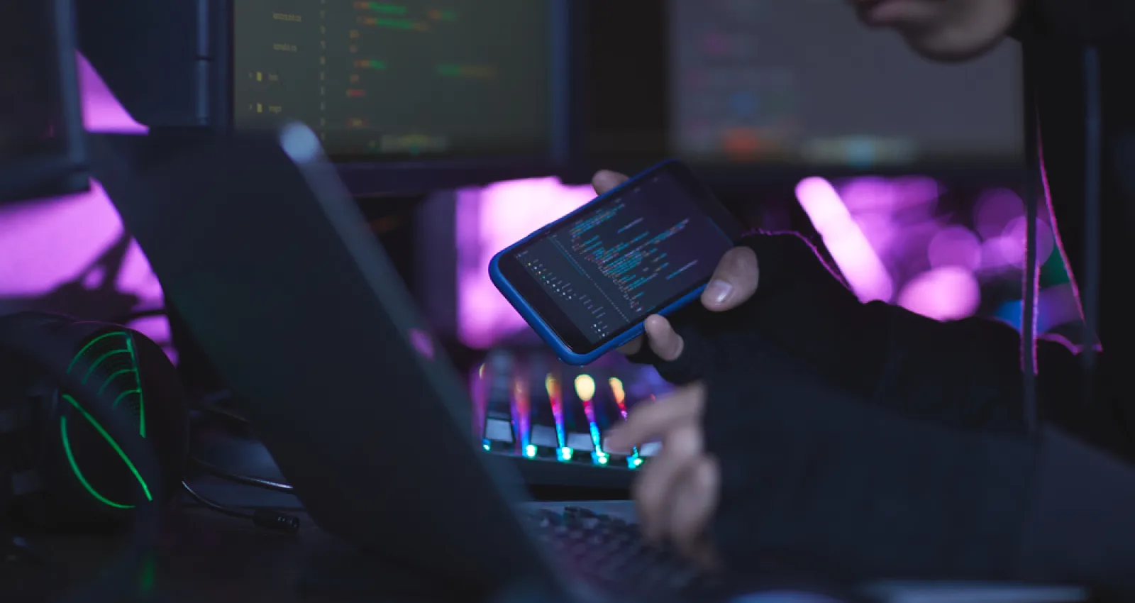 representación de hacker, con una computadora al frente y viendo datos en un celular a su costado derecho