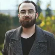 Foto de un hombre con barba y fondo verde con una gabardina gris mirando la cámara