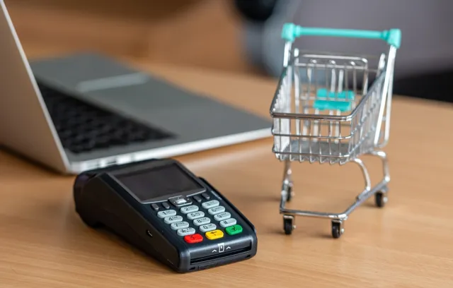 Foto de un datafono con un computador al fondo y al costado un carrito de compras