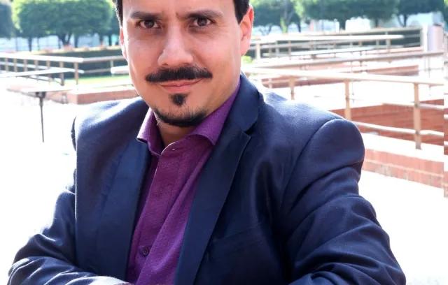 Foto de un hombre blanco con bigote mirando a la cámara, utiliza blazer azul y camiseta vinotinto