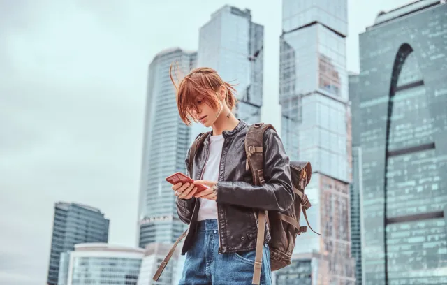 Foto de una mujer con chaqueta roja mirando un celular y unos edificios de fondo