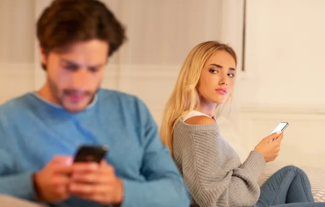 Foto de una mujer con un celular en la mano mirando de forma sospechosa a su pareja que habla por celular