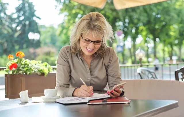 Foto de una mujer rubia anotando algo en un cuaderno con un fondo de arboles