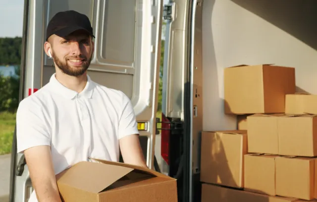 Foto de un un hombre con camiseta blanca y gorra negra alzando una caja sonriendo a la cámara