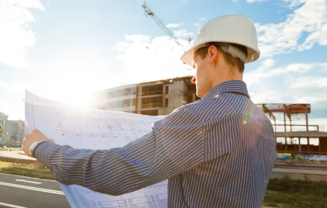 Fotografía de un constructor con camiseta azul sosteniendo en sus manos un plano de construcción y una edificación de fondo