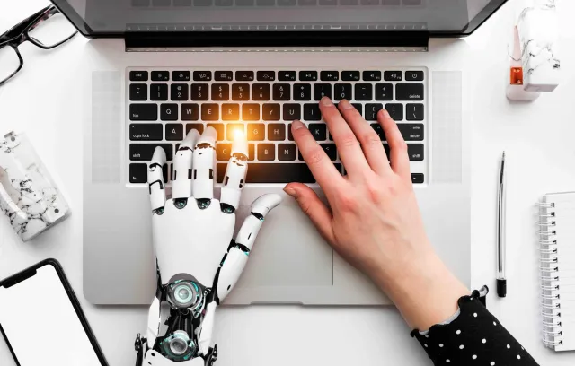 Foto de una mano biónica y humana escribiendo sobre un pc