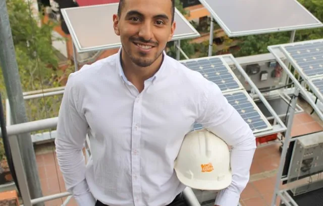 Foto de una persona con un casco posando frente a la cámara con unos paneles solares de fondo