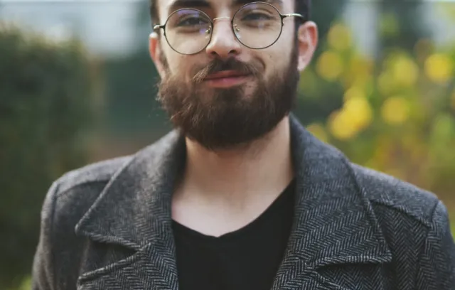 Foto de un hombre con barba y fondo verde con una gabardina gris mirando la cámara