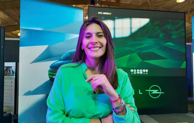 Una mujer con los brazos cruzados mirando a la cámara con una pantalla grande que en el fondo dice Opel