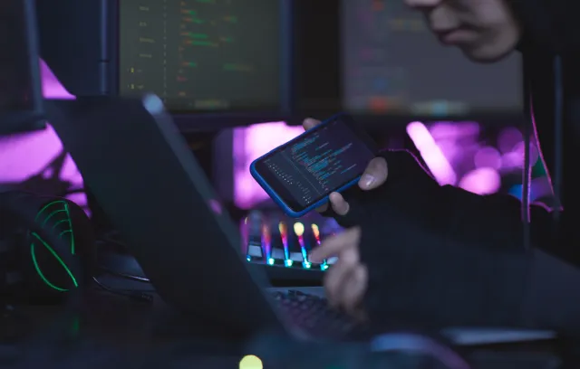 representación de hacker, con una computadora al frente y viendo datos en un celular a su costado derecho